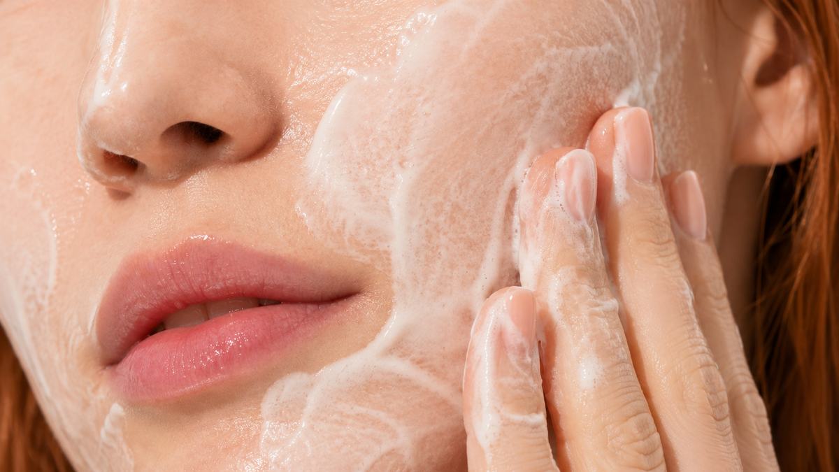 Close-up of fine cleansing foam being worked gently across the cheek with two fingertips