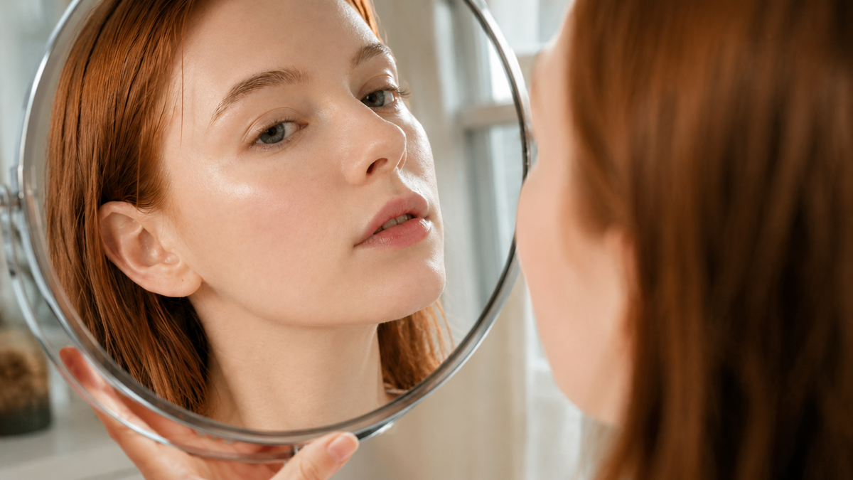 Woman closely checking her skin in a round hand mirror in soft natural light